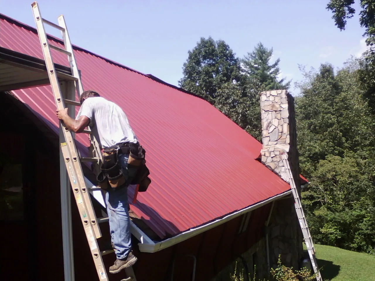 Roofer on a ladder beside a red metal roof and stone chimney — Soffit & Fascia Repair by Roofing Brothers in Counce
